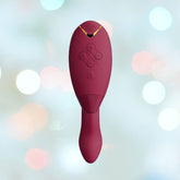 The Womanizer Duo 2 Rabbit Air Pulse Vibrator in Bordeaux is a maroon handheld electronic device with a curved shape and buttons on its front. Enhanced by Pleasure Air Technology for double stimulation, it promises an unparalleled experience. The background is softly blurred with pastel-colored lights, creating a dreamy atmosphere.